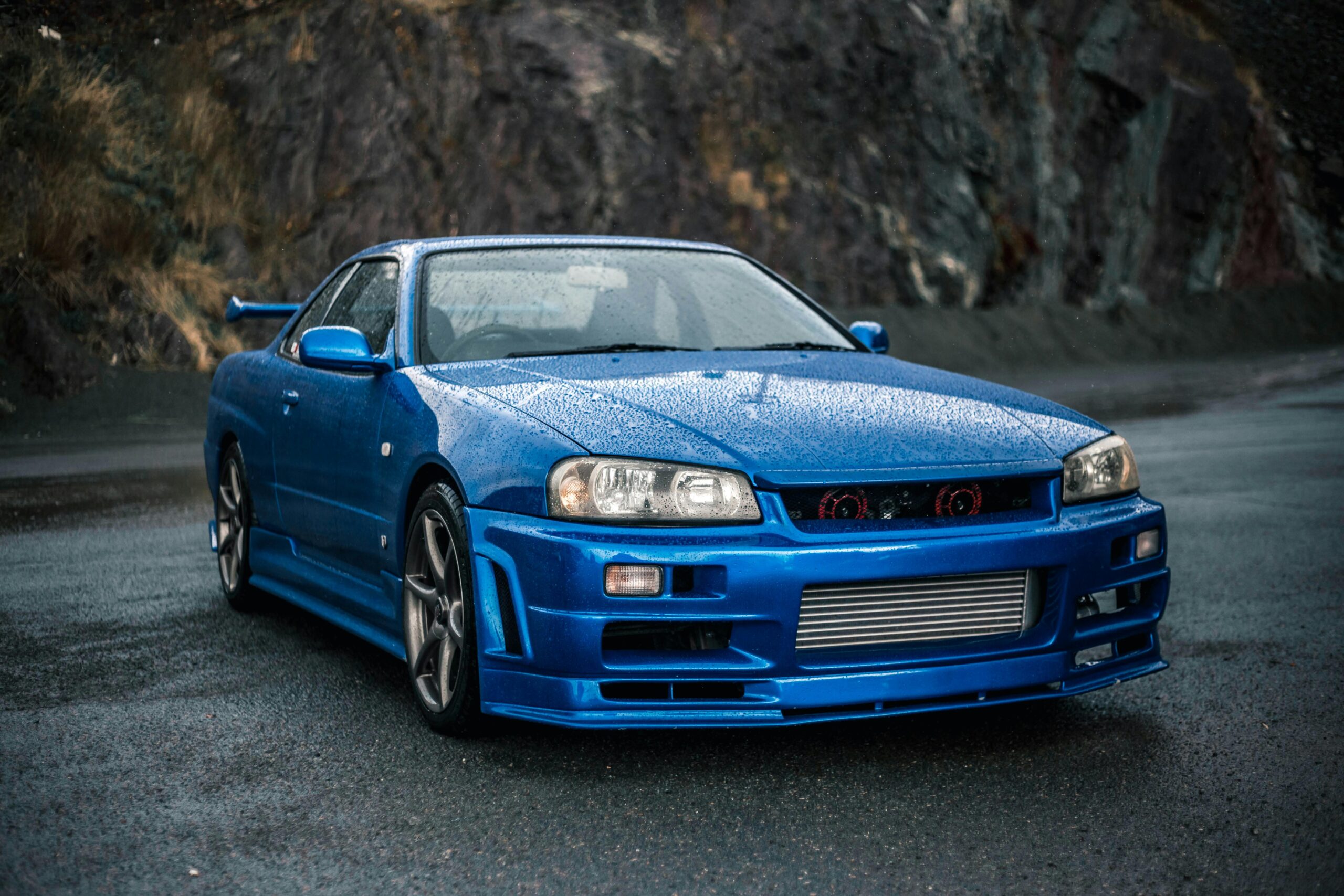 A striking blue Nissan Skyline R34 sports car parked on a wet, mountainous road with a misty background.